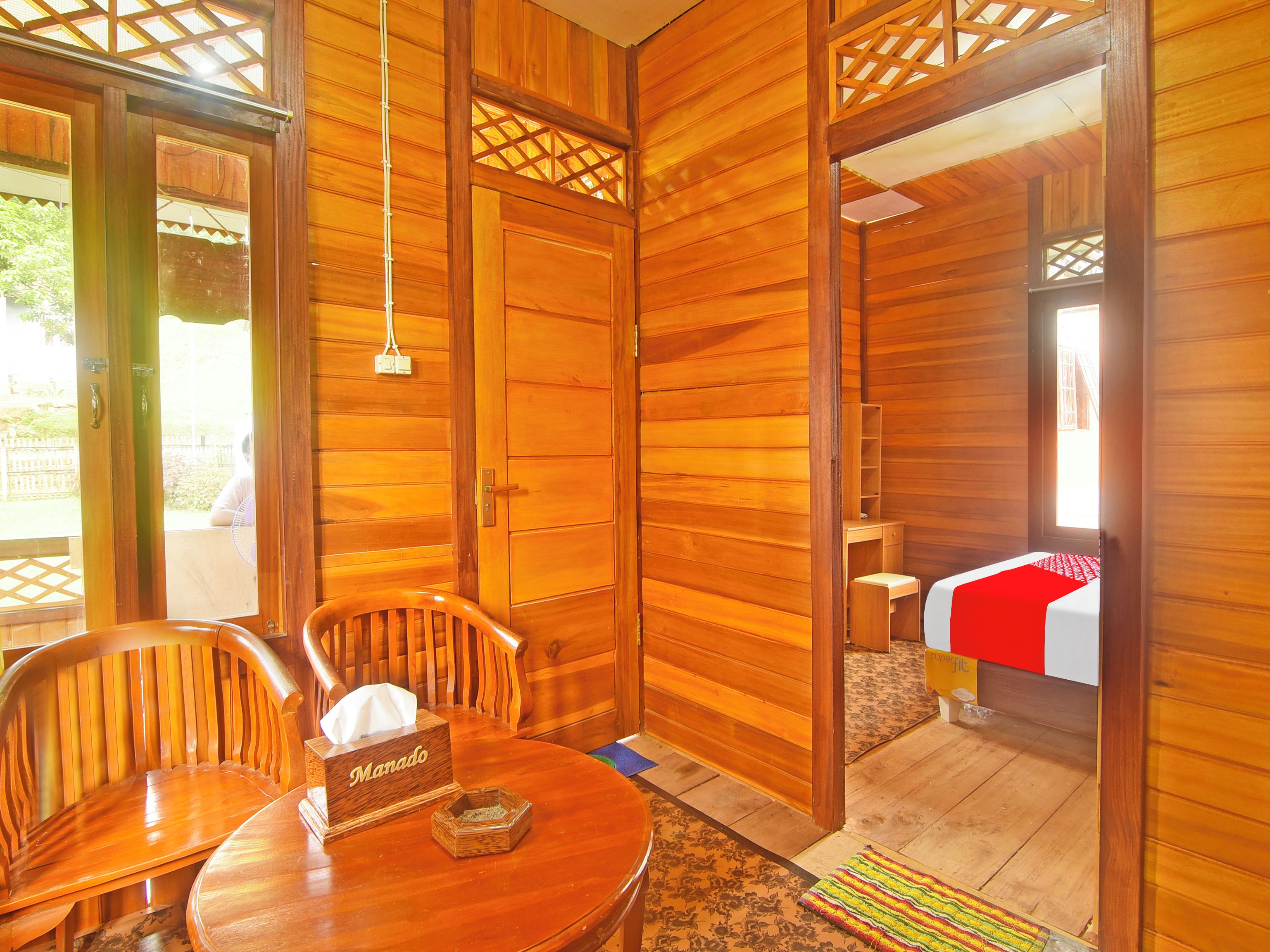 A wooden room with a round table, two chairs, a bedside table with a red and white bedspread, and a small wooden box labeled "Manado".