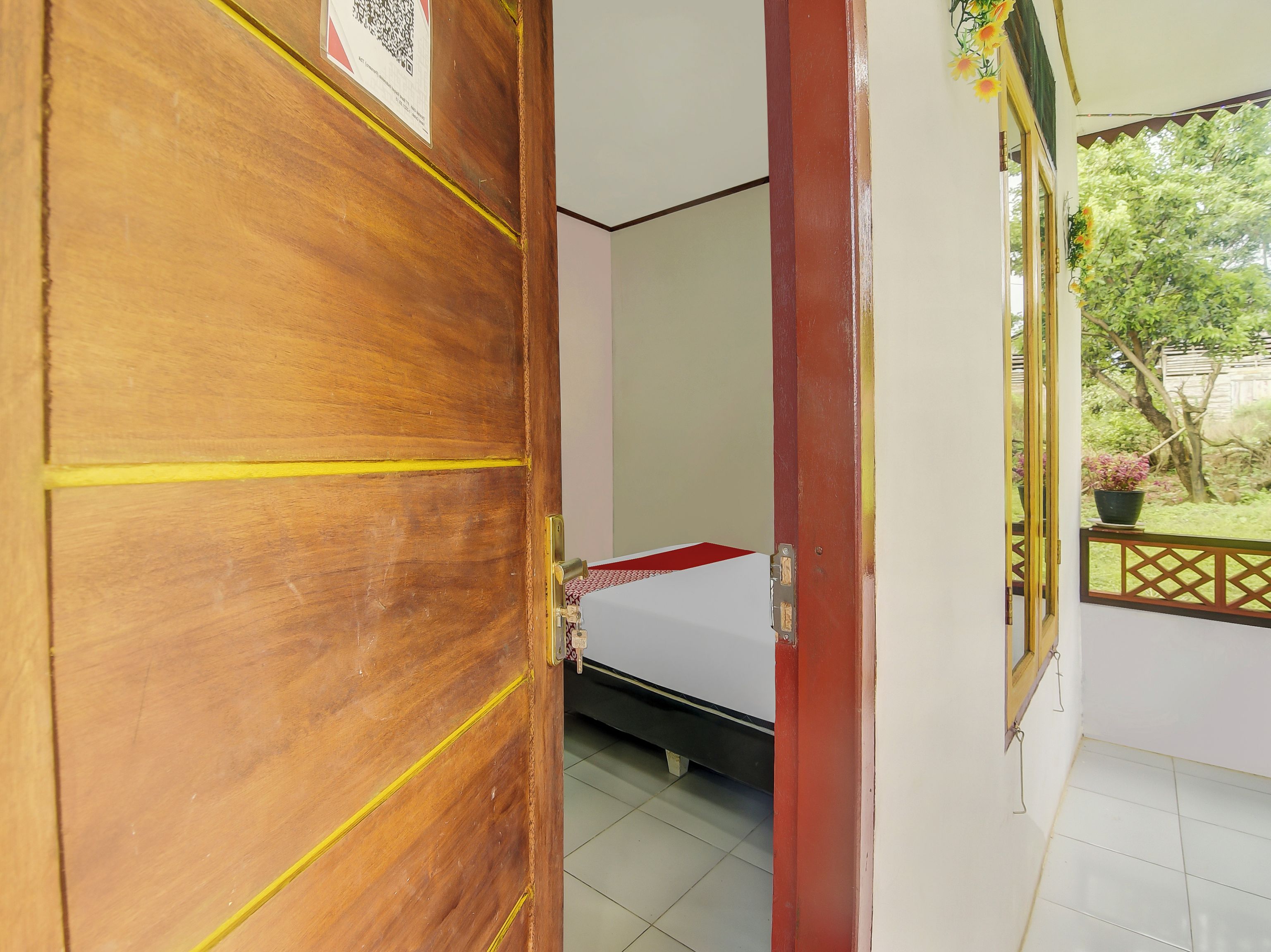 A wooden door with yellow markings, a bed with a white mattress and red accent, and a window with a view of greenery.