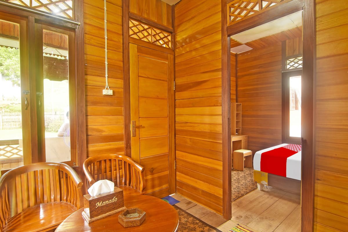 A wooden room with a round table, two chairs, a bedside table with a red and white bedspread, and a small wooden box labeled "Manado".