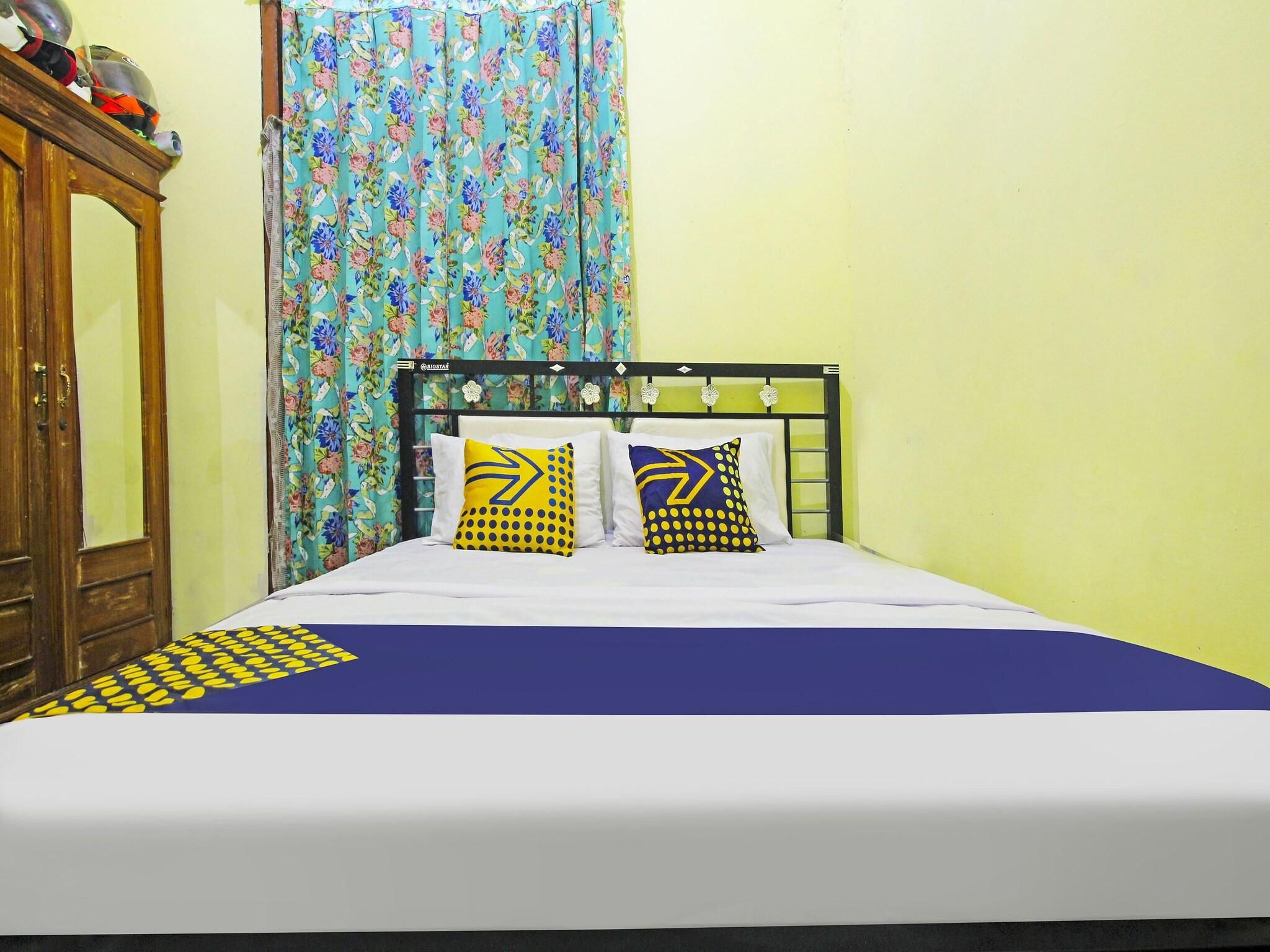 A bed with a blue bedspread and two pillows with yellow and blue patterns, a wooden wardrobe, and a floral curtain.