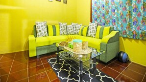A living room with a bright yellow wall, a green sofa with patterned cushions, a glass coffee table with decorative items, and a floral curtain. - OYO Homes 91149 Desa Wisata Nangelasari (Nangelasari)