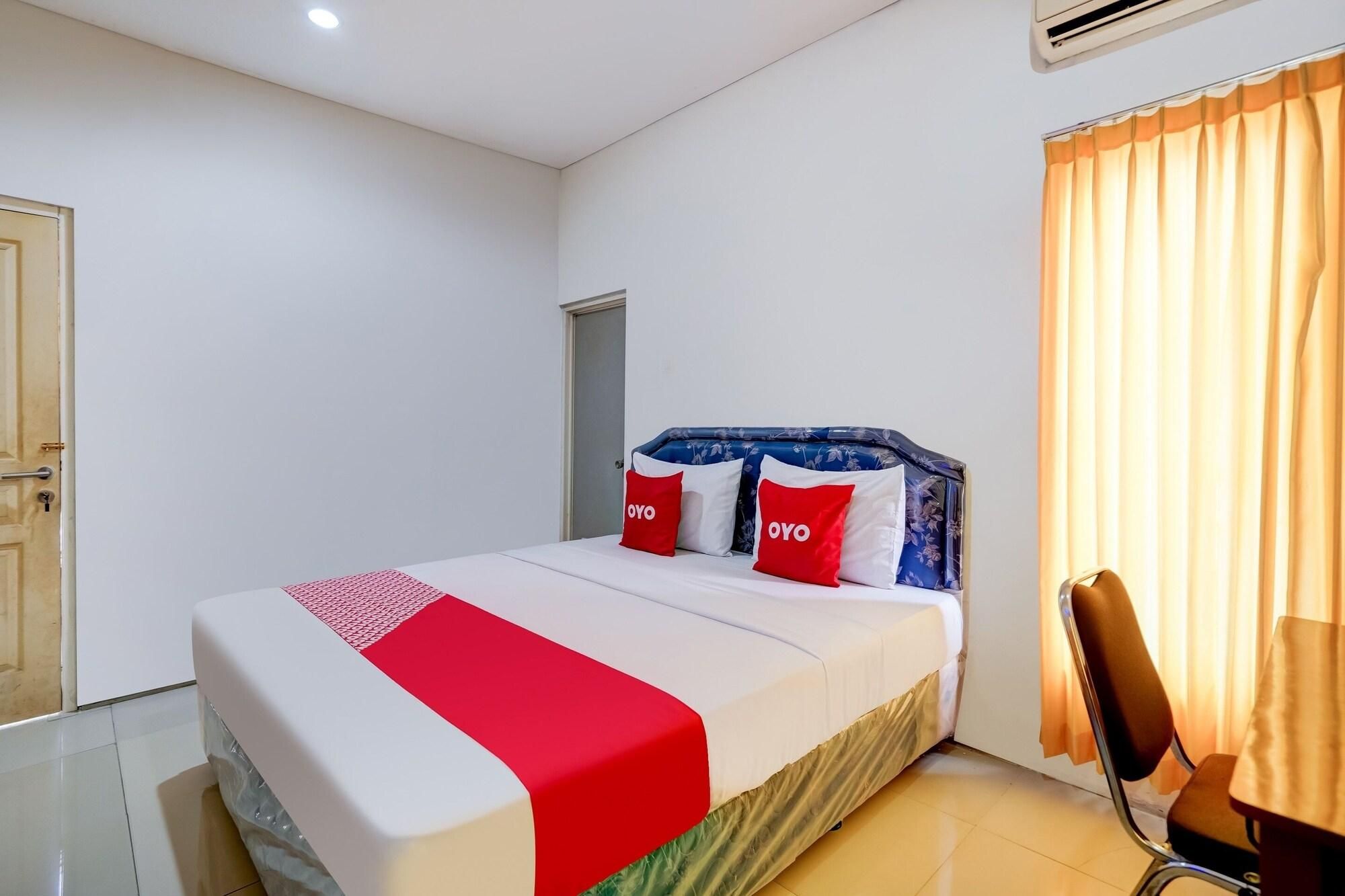 Uma cama de solteiro com edredom branco e vermelho, duas almofadas com a marca 'OYO', cabeceira azul e uma cadeira em quarto com paredes brancas e janela com cortinas.