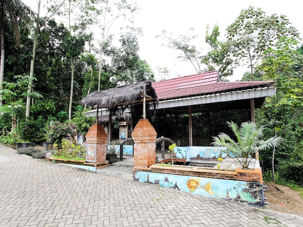 A rustic building with a thatched roof and brick pillars, surrounded by lush greenery and a paved entrance.
