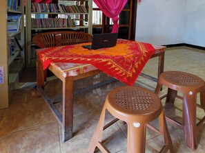 Una habitación con una mesa de madera, una computadora portátil, dos taburetes de plástico y una estantería llena de libros.