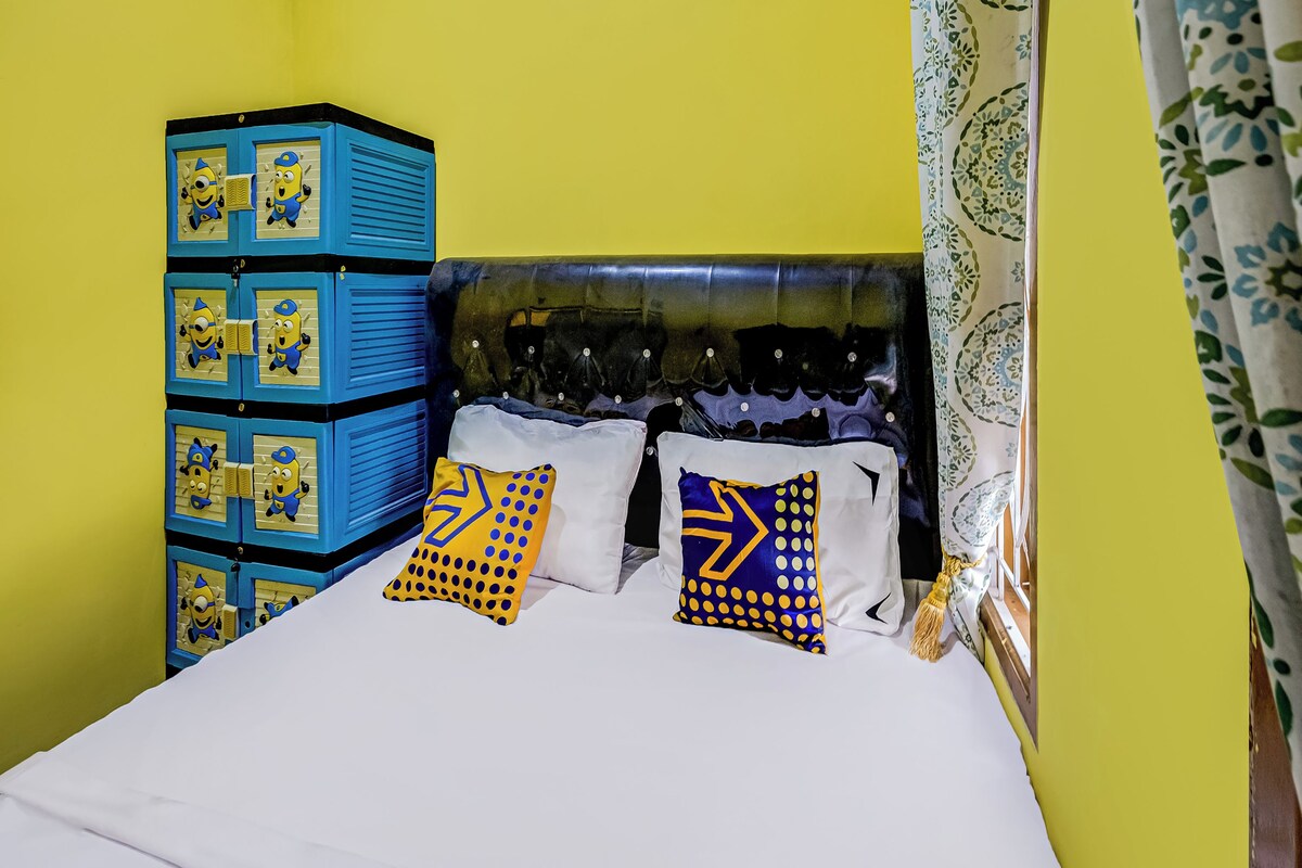 A bed with white bedding, two pillows with geometric patterns, a blue storage unit with Minion designs, and a tufted headboard.
