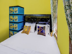 Un lit avec une literie blanche, deux oreillers à motifs géométriques, une unité de rangement bleue ornée de motifs de personnages de « Moi, moche et méchant », et une tête de lit capitonnée.