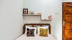 A bedroom with a wooden bed frame, white bedding, and two pillows with geometric patterns. A shelf above the bed holds a framed photo, a birthday card, and a stuffed toy.