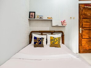 Un dormitorio con estructura de cama de madera, ropa de cama blanca y dos almohadas con diseños geométricos. Un estante sobre la cama sostiene una fotografía enmarcada, una tarjeta de cumpleaños y un peluche.