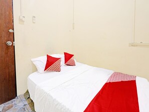 A single bed with white bedding and red accents, a wooden door, and a plain wall with a light switch.