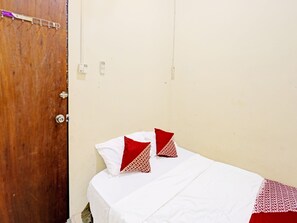 A small, simple room with a wooden door, a bed with white bedding and red patterned pillows, and a plain wall.