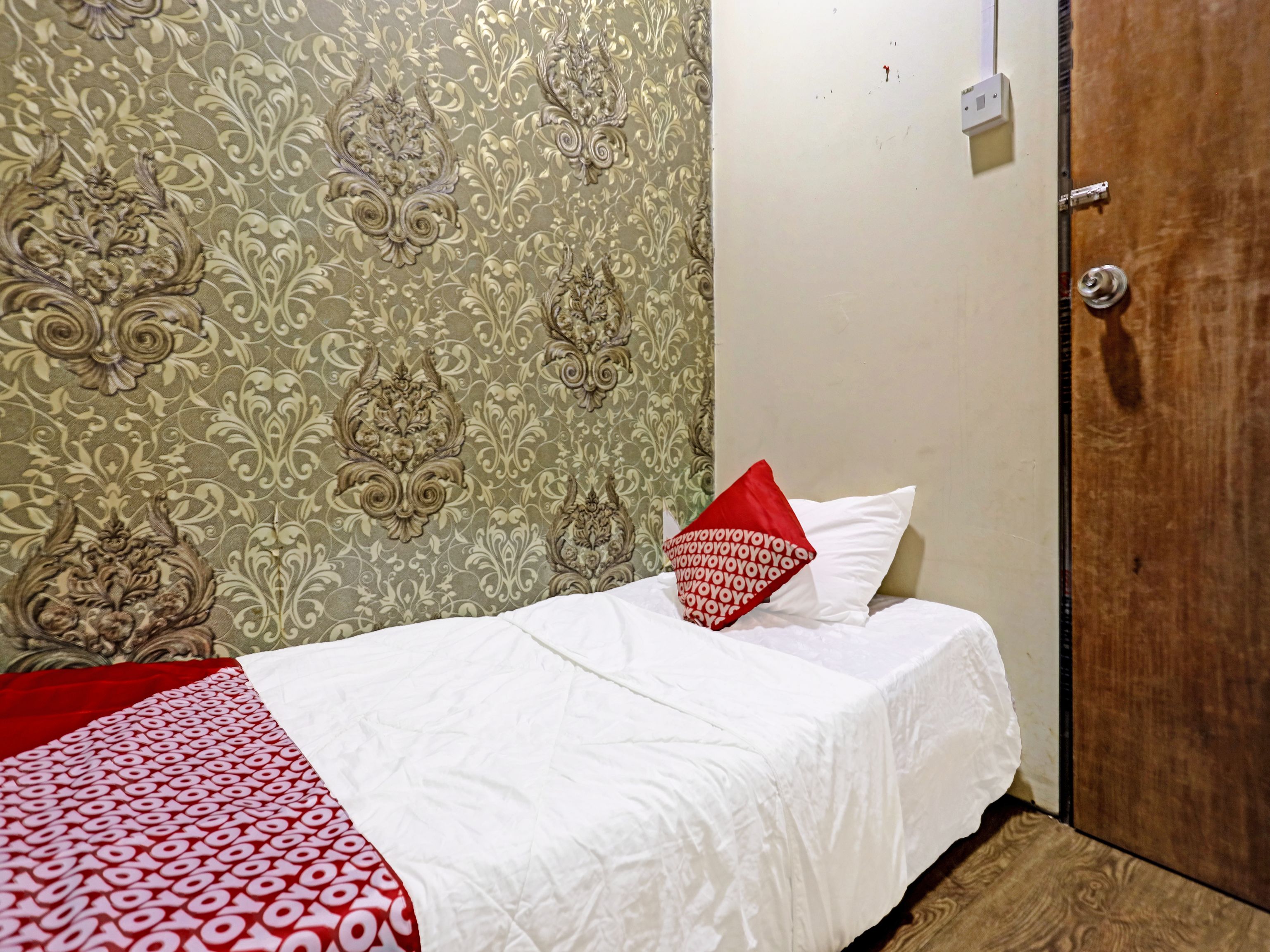 A single bed with a white bedspread and a red pillow, a wooden door, and patterned wallpaper.