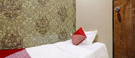 Una cama individual con una colcha blanca y un almohadón rojo, una puerta de madera y un papel tapiz estampado.