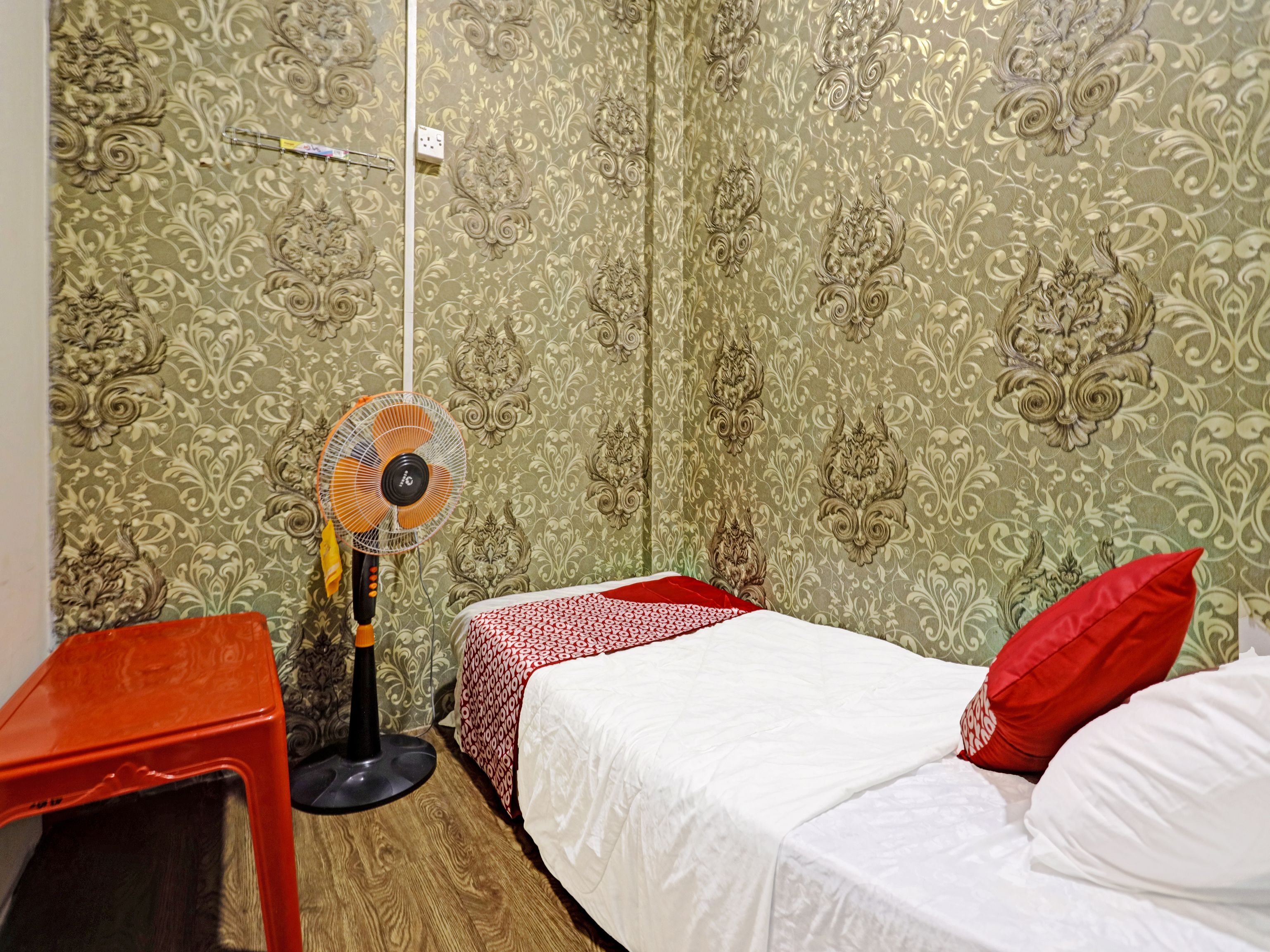 A small room with a single bed, a bedside table, a standing fan, and patterned wallpaper.