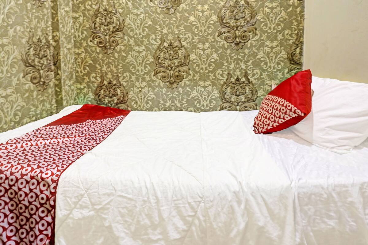 A neatly made bed with a white and red bedspread, a red pillow, and a green patterned curtain in the background.