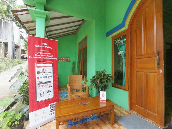 A green building with a wooden door, a red signboard, a wicker chair, a wooden table, and a potted plant.