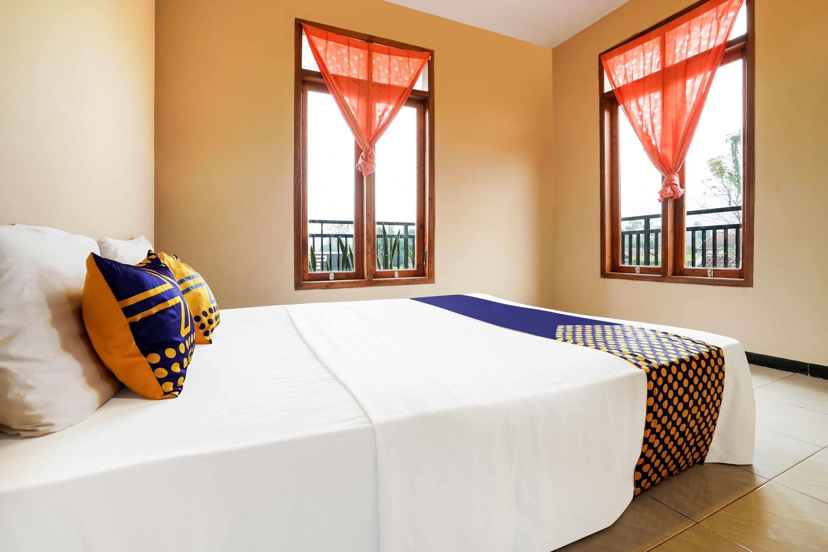 A hotel room with a bed, two windows with curtains, a balcony, and a table with a patterned tablecloth.