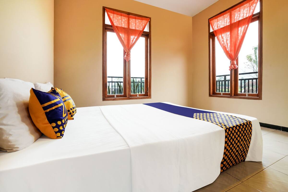 Ein Hotelzimmer mit Bett, zwei mit Vorhängen versehenen Fenstern, einem Balkon und einem Tisch mit gemustertem Tischläufer.