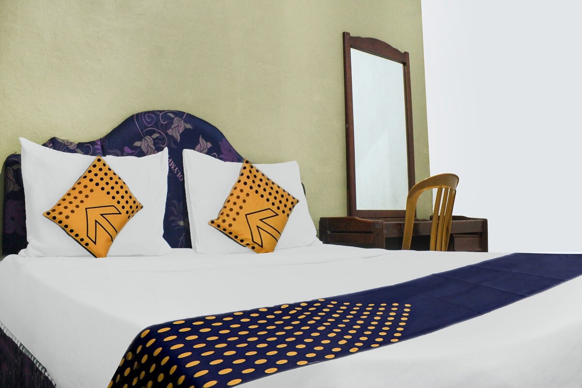 Un letto con lenzuola bianche e due cuscini a motivo geometrico, una coperta azzurra a pois, una sedia di legno e uno specchio appeso al muro.