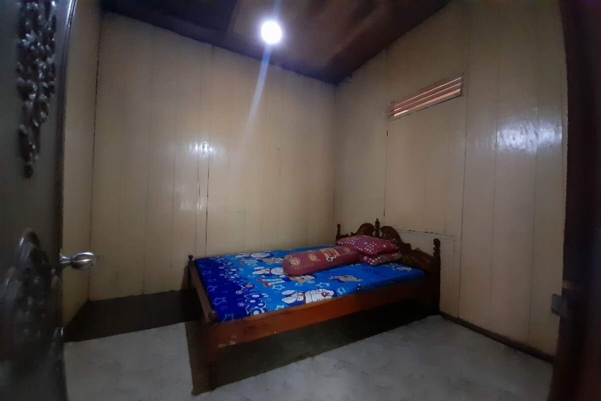 A small, single-bed room with a wooden headboard, a blue bedspread with cartoon characters, and a wooden door with a metal handle.