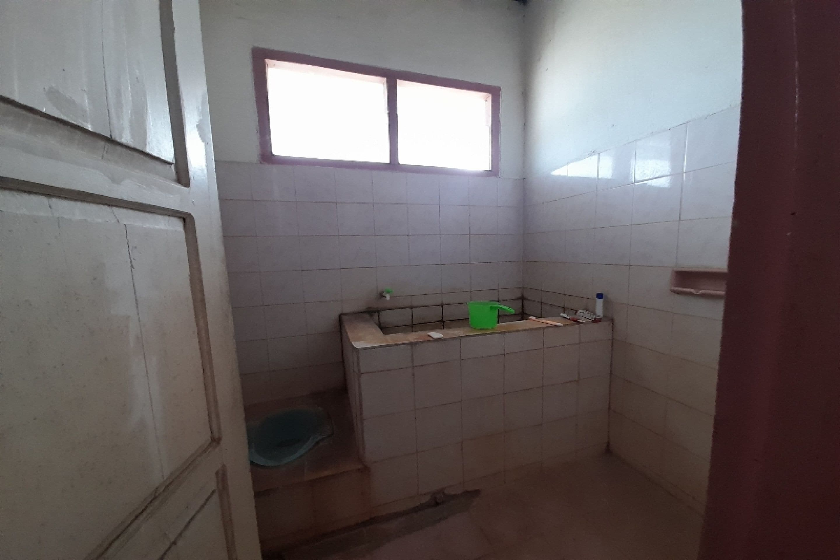 A small, tiled bathroom with a window, a green bucket, and a blue basin.