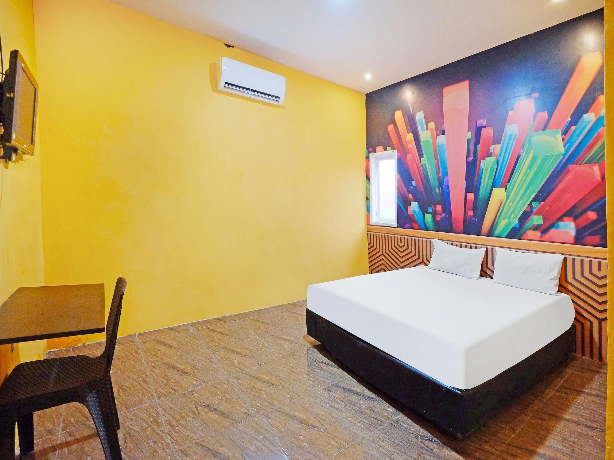 Quarto de hotel com cama, escrivaninha, cadeira e um quadro abstrato colorido na parede.