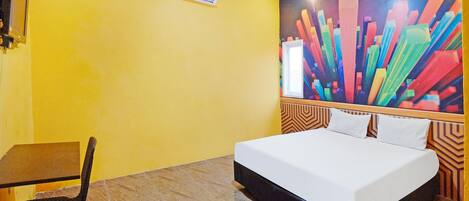 Quarto de hotel com cama, escrivaninha, cadeira e um quadro abstrato colorido na parede.