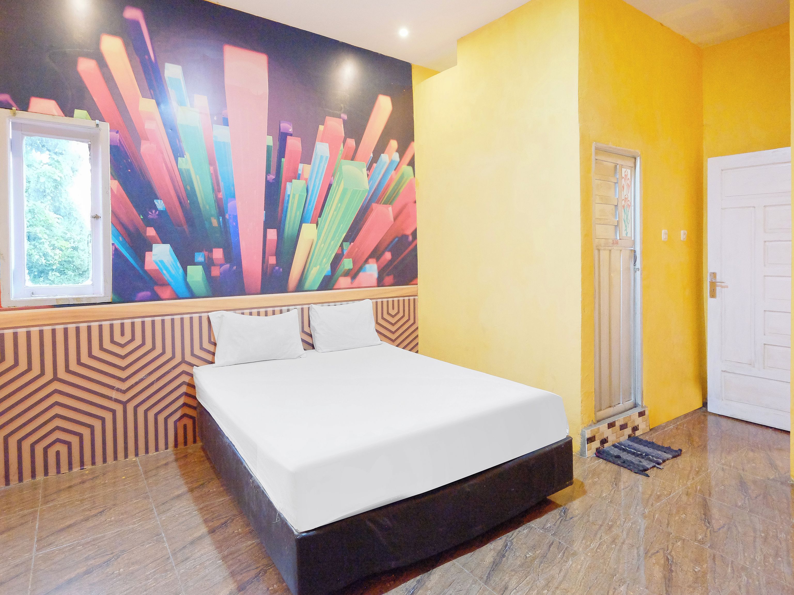 Habitación de hotel con cama, un mural abstracto de colores, una ventana que da a zonas verdes y una puerta.