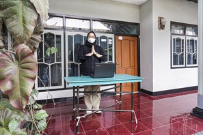 Una persona de pie detrás de una mesa con una computadora portátil, en una habitación con piso de baldosas y ventanas amplias.