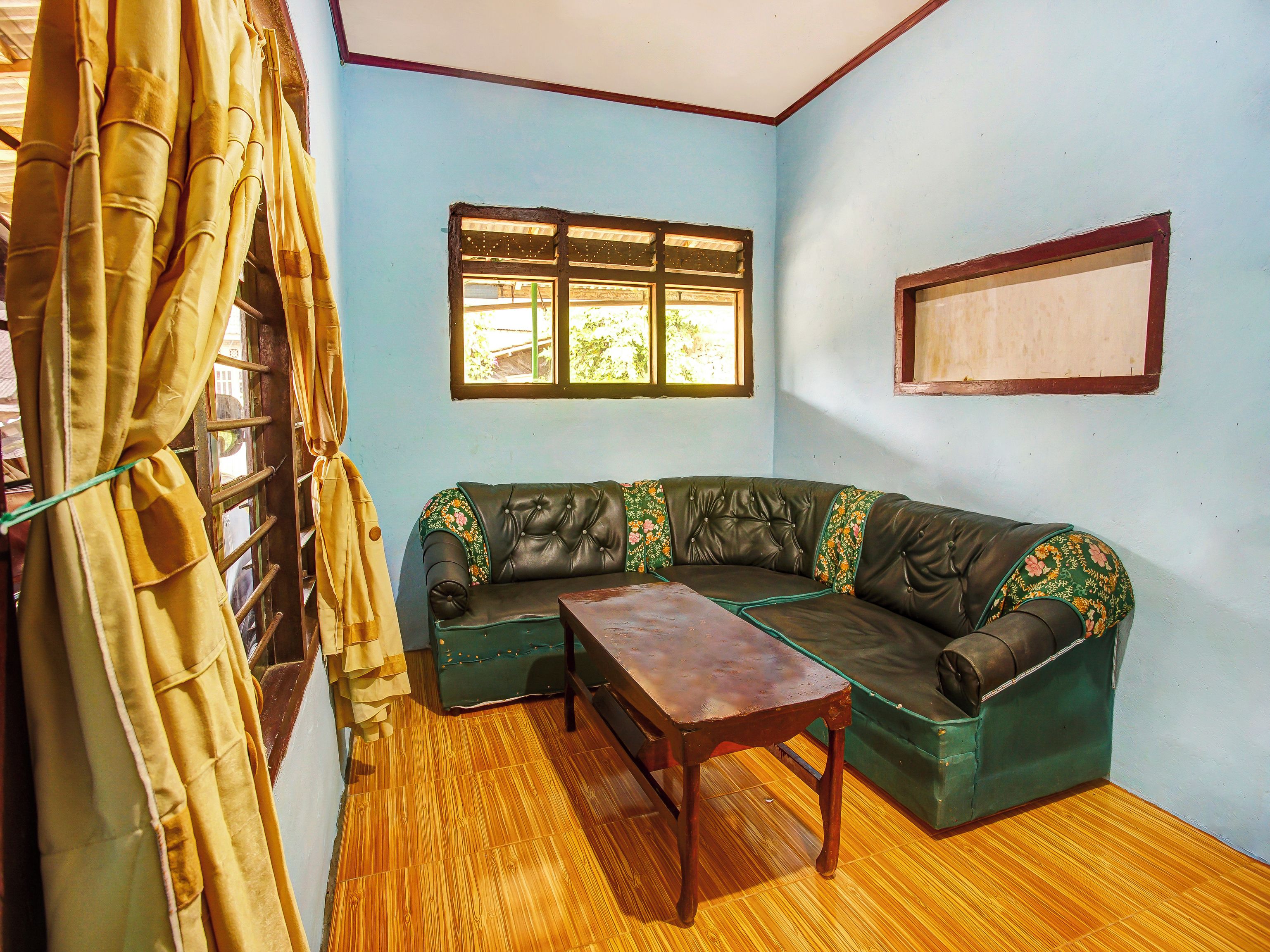A room with a wooden floor, a green sofa with floral cushions, a wooden table, and a window with a view.
