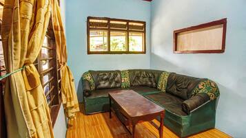 A room with a wooden floor, a green sofa with floral cushions, a wooden table, and a window with a view.