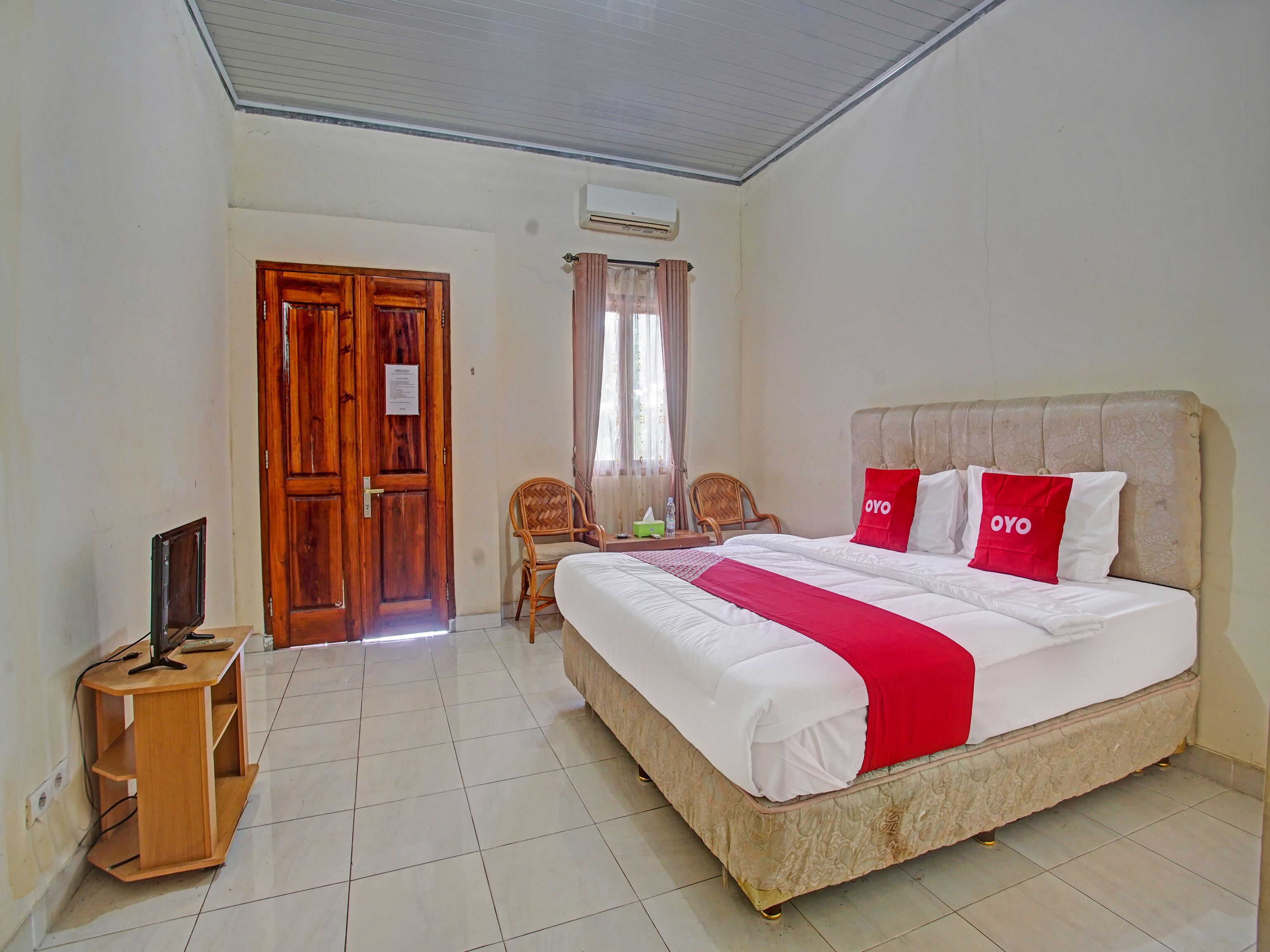 A hotel room with a large bed, two chairs, a wooden desk, and an air conditioning unit.