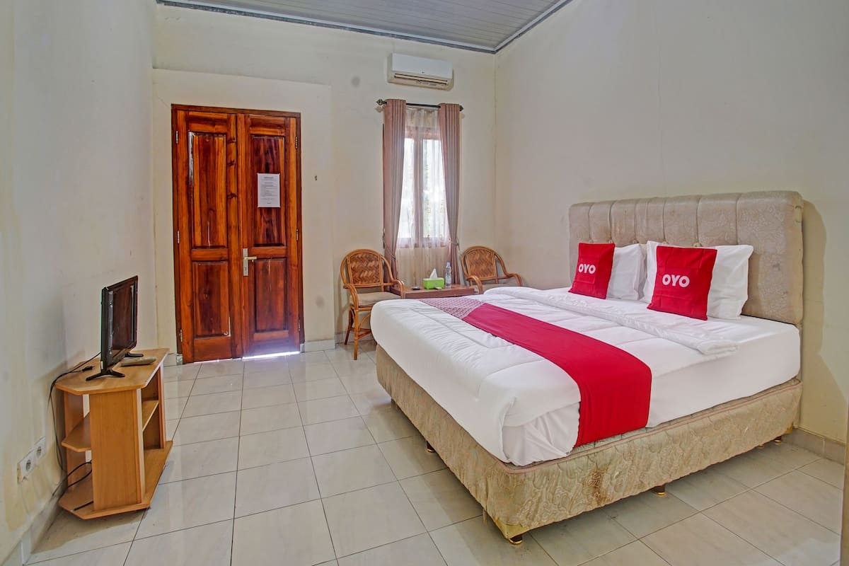 A hotel room with a large bed, two chairs, a wooden desk, and an air conditioning unit.