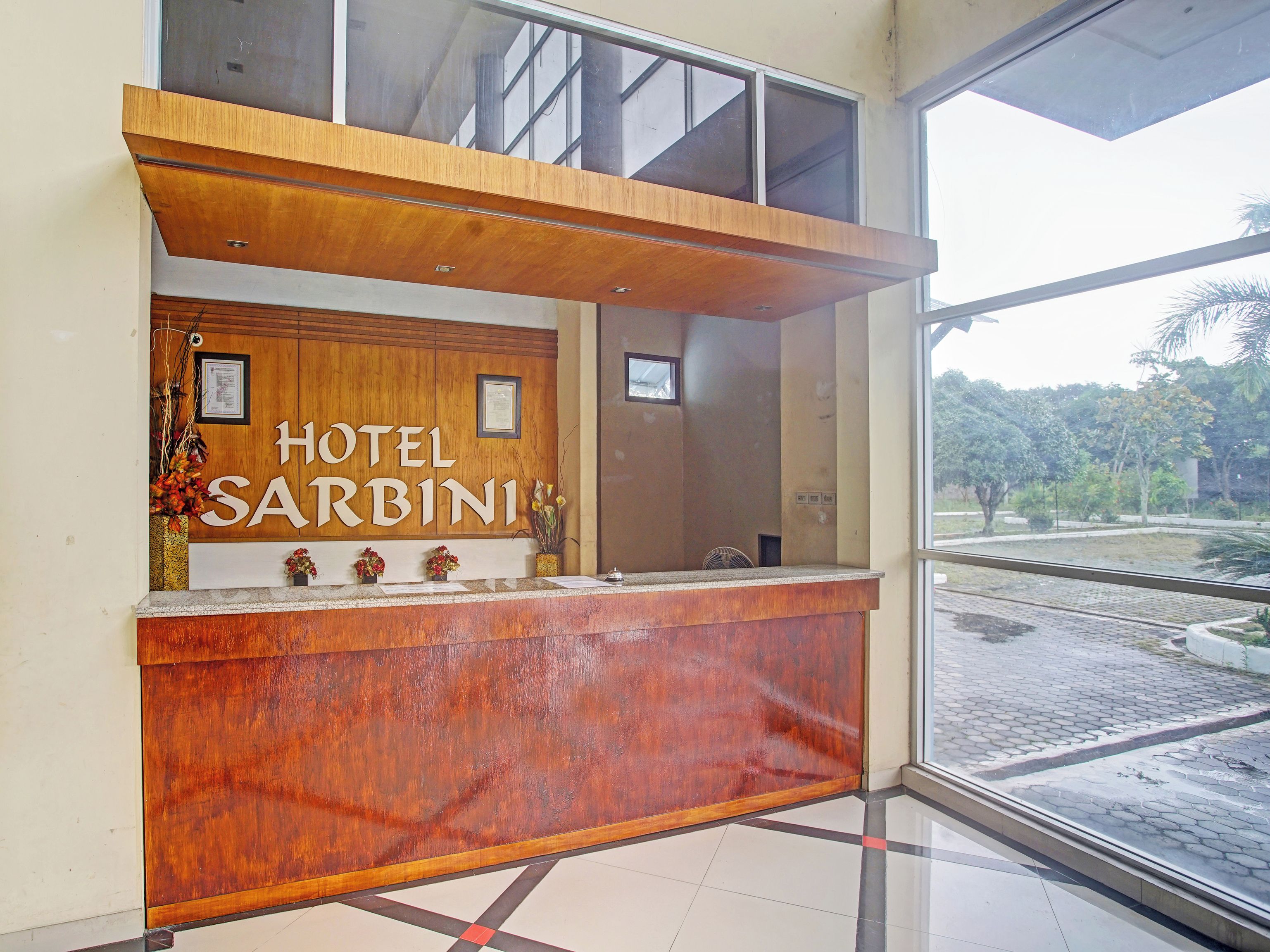 A reception desk with a wooden finish, a sign reading "HOTEL SARBINI", and a view of a landscaped area outside.