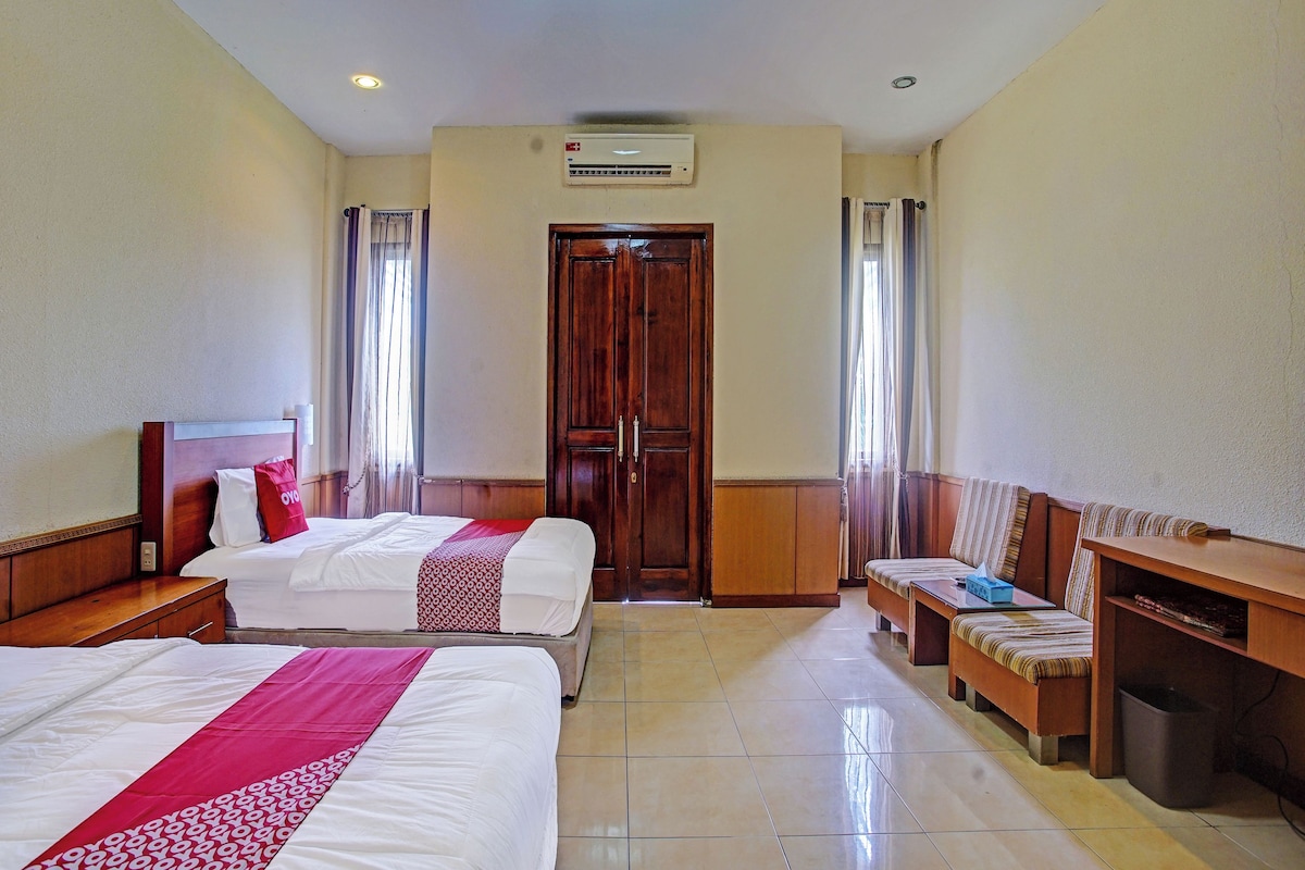 A hotel room with two beds, a wooden wardrobe, a bench, a desk, and a trash bin.