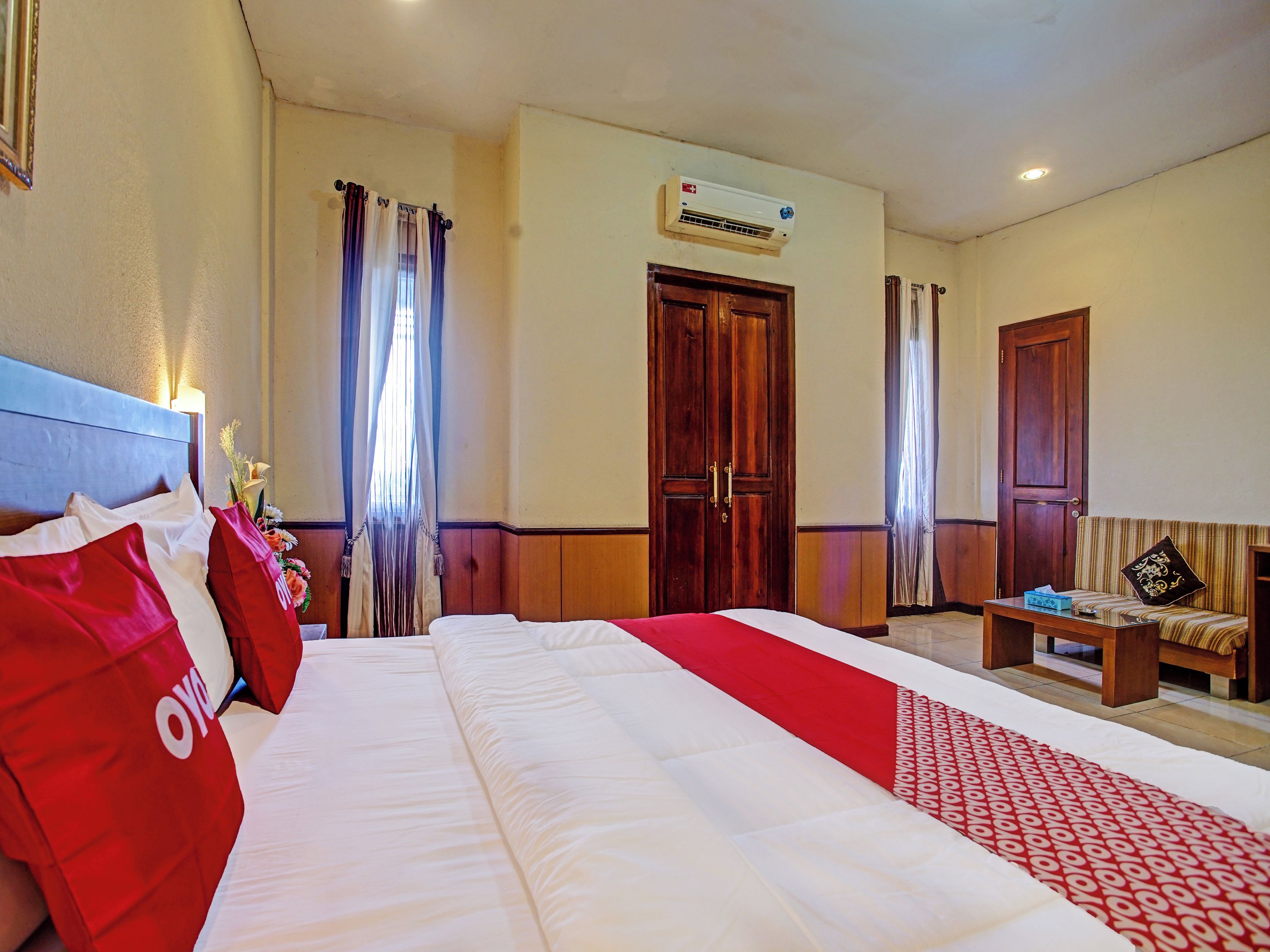 A hotel room with a large bed, a wooden wardrobe, a small table, and an air conditioning unit on the wall.