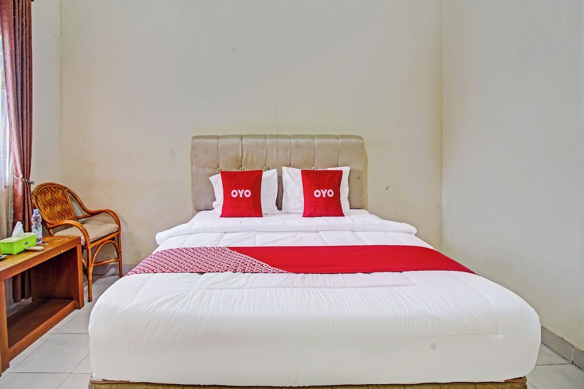 A neatly made bed with white and red bedding, a wooden chair, and a small table with a bottle and tissue box.