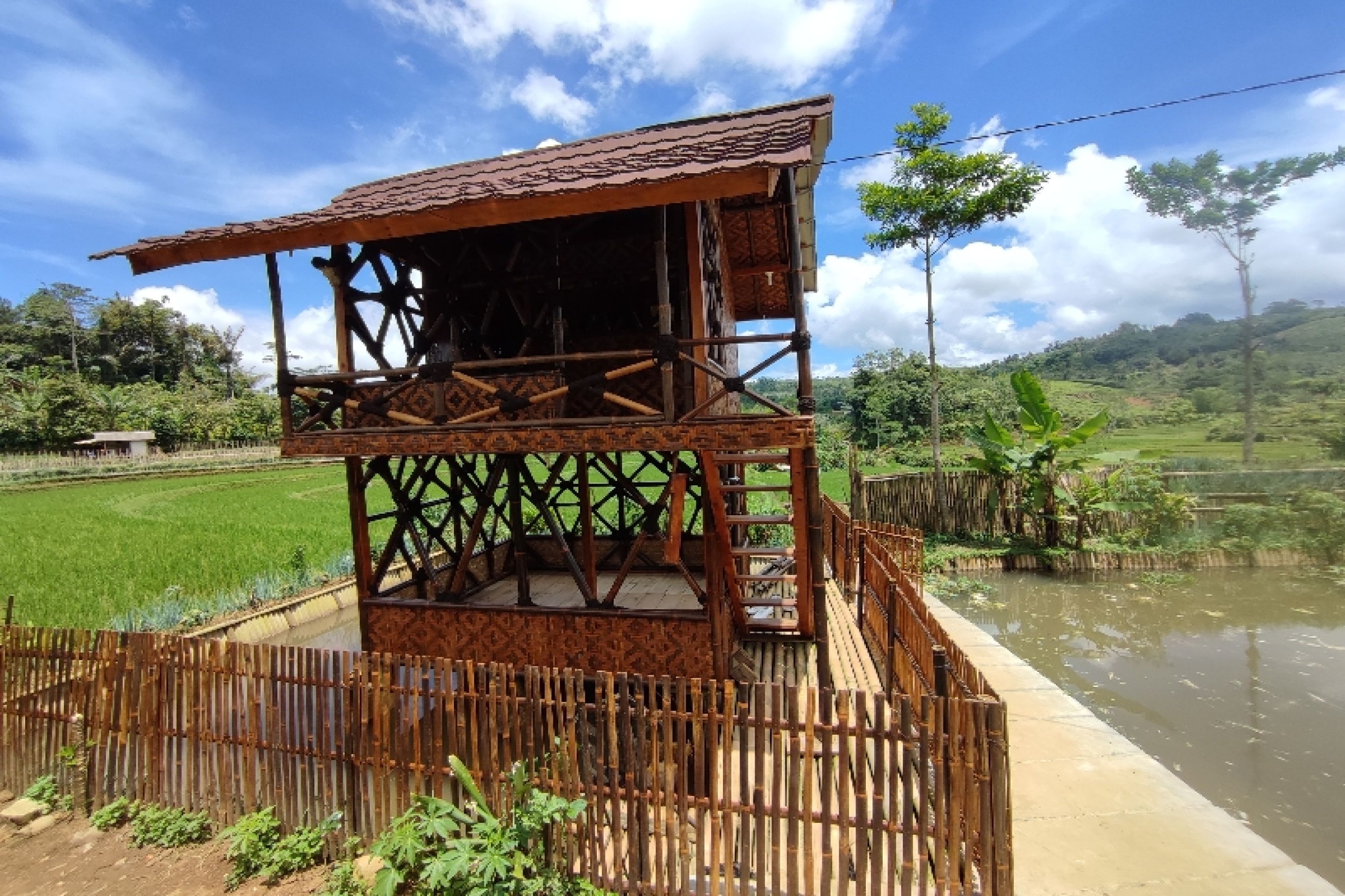 A rustic wooden structure with a balcony, surrounded by lush greenery and a water body.