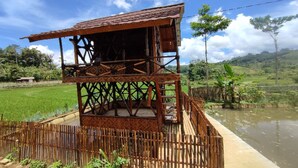 A rustic wooden structure with a balcony, surrounded by lush greenery and a water body.