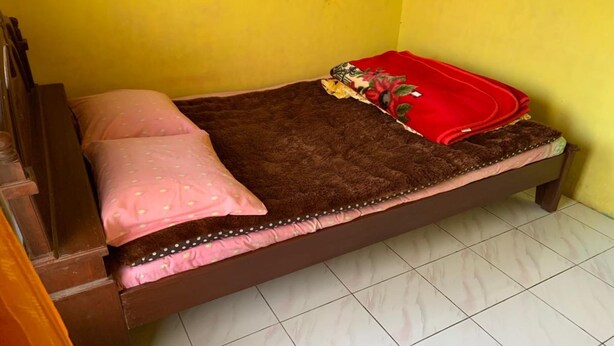 A single bed with a brown mattress, pink and white patterned pillows, and a red blanket.