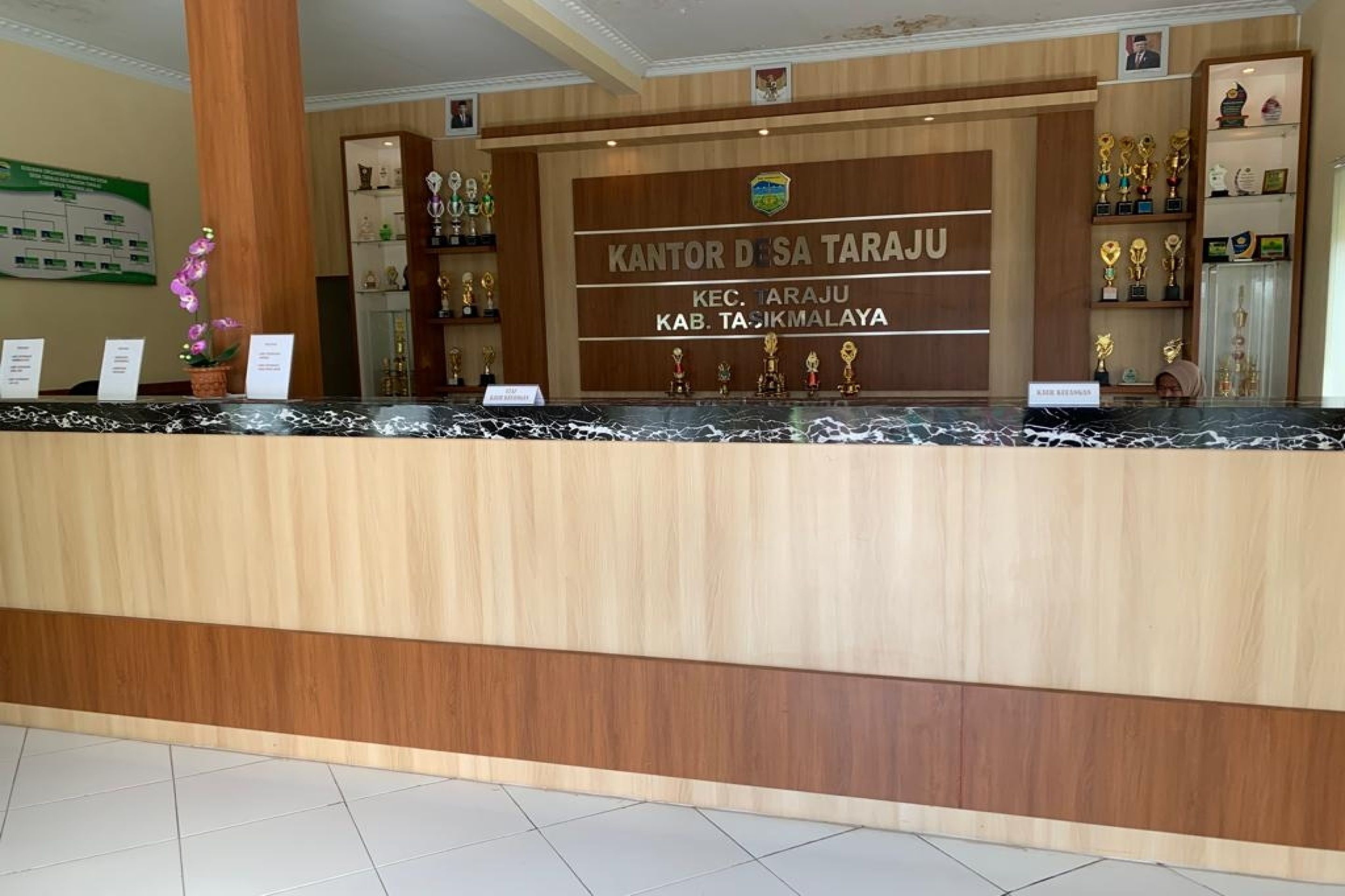 A reception desk with a wooden finish and a marble top. Behind the desk, there is a sign with the text "KANTOR DESA TARAU" and "KEC. TARAUU KAB. TASIKMALAYA".