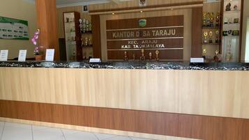 A reception desk with a wooden finish and a marble top. Behind the desk, there is a sign with the text "KANTOR DESA TARAU" and "KEC. TARAUU KAB. TASIKMALAYA".