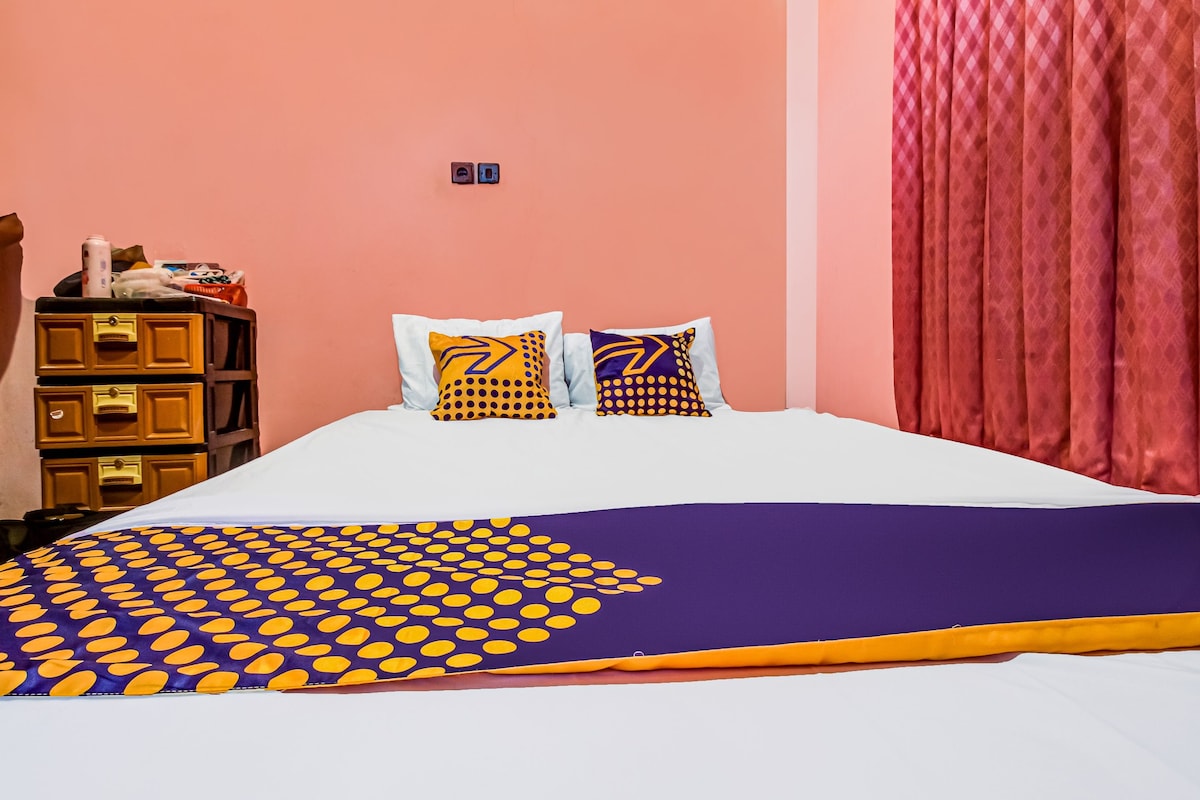 Uma cama com colcha estampada, duas almofadas com desenho geométrico, um penteadeira de madeira e uma cortina vermelha.