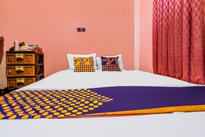 Uma cama com colcha estampada, duas almofadas com desenho geométrico, um penteadeira de madeira e uma cortina vermelha.