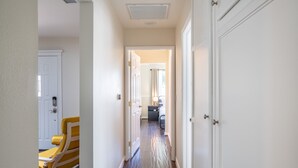 A hallway with a yellow chair, white doors, and a view into a room with a bed and a lamp.