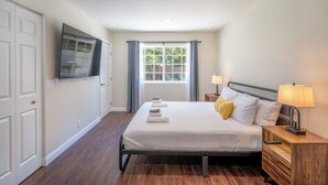 A bedroom with a bed, a nightstand, a TV, a window with curtains, and a door.
