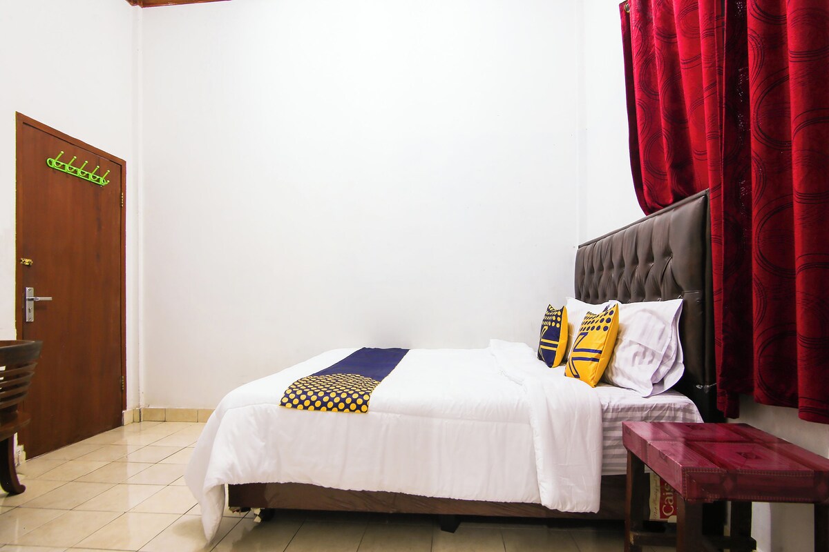 Uma cama bem arrumada com edredom branco e duas almofadas decorativas, uma porta de madeira com um porta-roupas verde e uma cortina vermelha.
