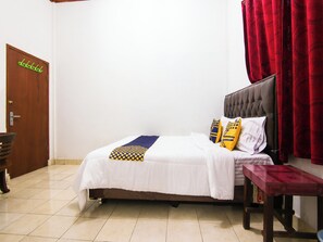 Uma cama bem arrumada com edredom branco e duas almofadas decorativas, uma porta de madeira com um porta-roupas verde e uma cortina vermelha.