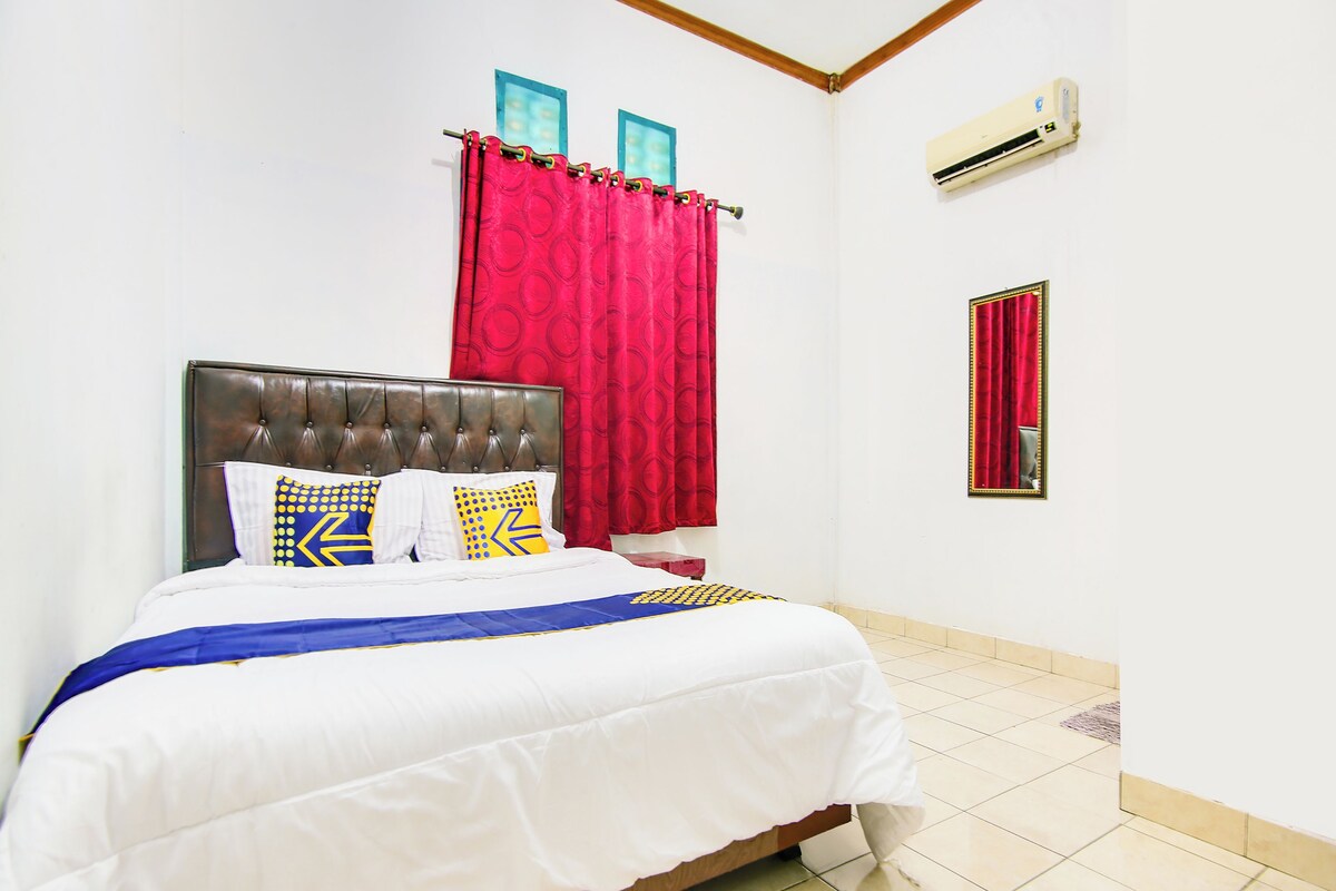 A neatly made bed with a headboard, a red curtain, a wall-mounted air conditioner, and a framed mirror.