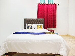 Cama bem arrumada com colcha branca e azul, duas almofadas com padrões geométricos, cabeceira estofada marrom escura e cortina vermelha.