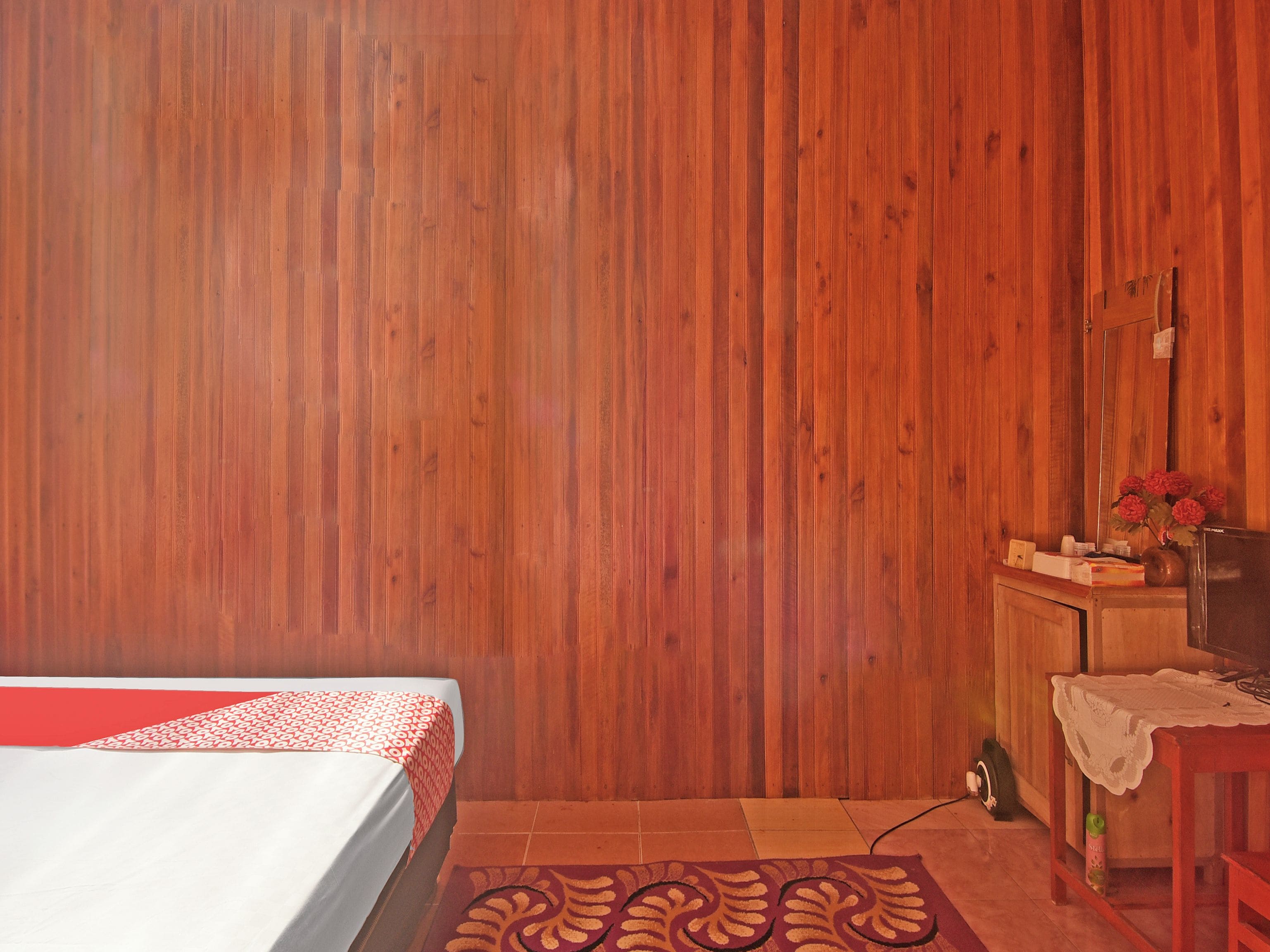 A room with a wooden wall, a bed with a red and white bedspread, a small table with a vase of flowers, and a mirror on the wall.